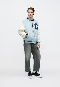 Φως μπλε varsity μπουφάν με κρεμ μανίκια, διαθέτει μπλε ραφτό γράμμα "C", ριμπ μανσέτες και γκρι τζιν παντελόνι με μαύρα αθλητικά παπούτσια.