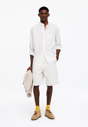 Man draagt een wit shirt met lange mouwen en een korte broek, houdt een beige jas vast, draagt gele sokken en lichtbruine suède schoenen, staand tegen een witte achtergrond.