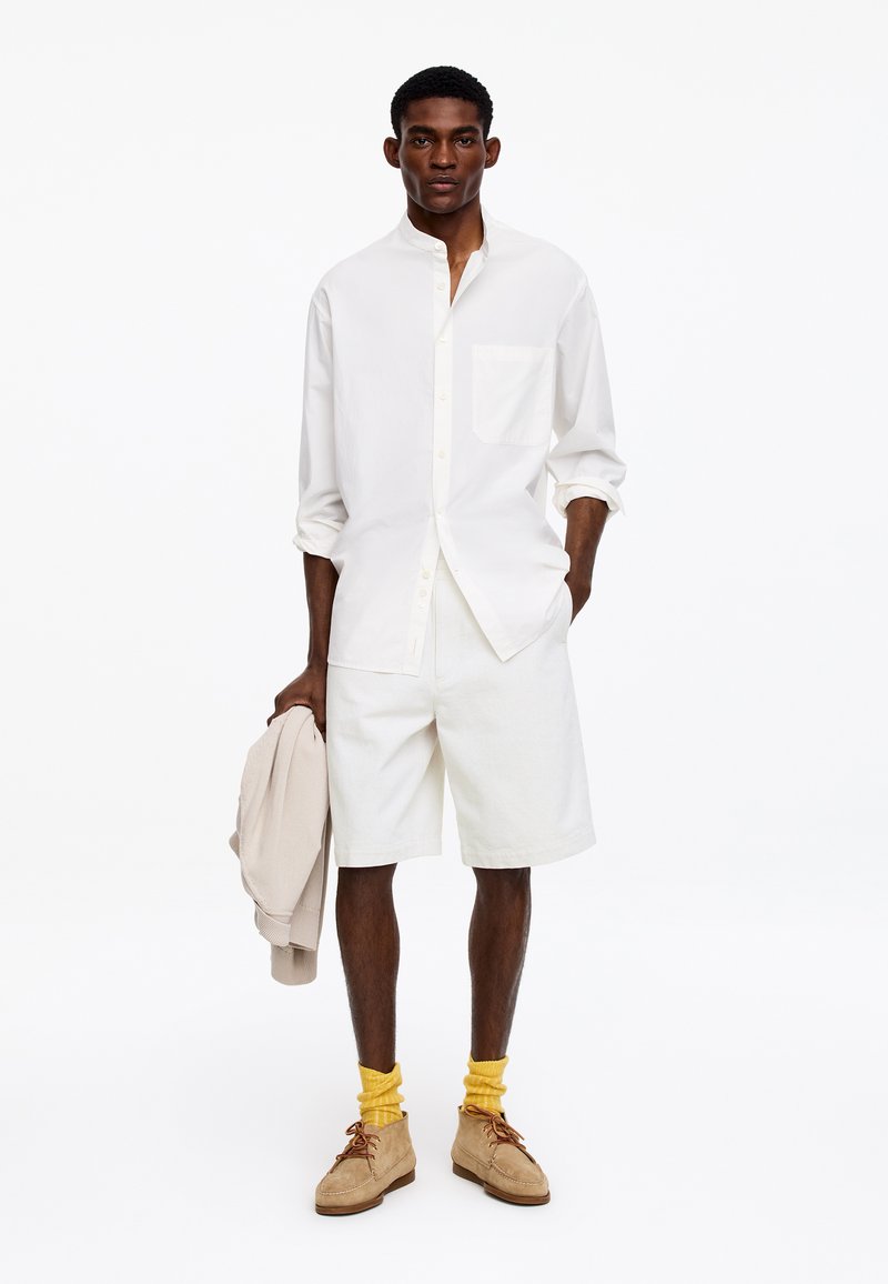 Homme portant une chemise blanche à manches longues et un short, tenant une veste beige, avec des chaussettes jaunes et des chaussures en suède beige, debout sur un fond blanc.