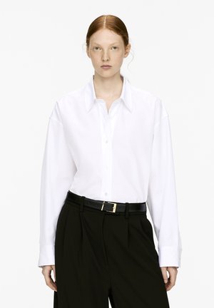 Junge Person mit rotem Haar, die ein weißes Hemd mit Knöpfen, schwarze Hosen und einen schwarzen Gürtel trägt, steht vor einem schlichten weißen Hintergrund.