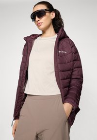 Maroon puffer jacket with a quilted design, paired with a light beige textured top and beige pants. Model wearing oversized black sunglasses.