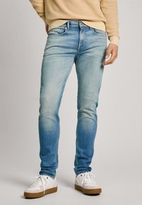 Jeans de mezclilla azul claro con corte slim, que presentan detalles de desgaste y cinco bolsillos, combinados con zapatillas blancas y un sudadera beige.