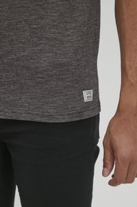 Camiseta gris de manga corta con rayas horizontales, que presenta una pequeña etiqueta blanca en el dobladillo, combinada con pantalones negros.