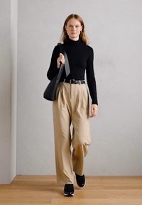 Un col roulé noir, un pantalon beige taille haute avec des plis, et des baskets noires. Porte un sac à main noir texturé. Fond neutre.