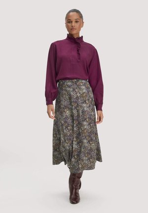 Frau trägt eine langärmelige lila Bluse mit Rüschendetails, einen geblümten Midirock und braune kniehohe Stiefel vor einem einfarbigen Hintergrund.