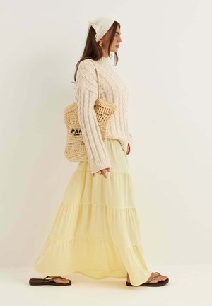 Kobieta idąca w kremowym swetrze z warkoczowym splotem, żółtej wielowarstwowej spódnicy maxi, dzianej opasce na głowę, brązowych sandałach i niosąca beżową plecioną torbę na ramię.