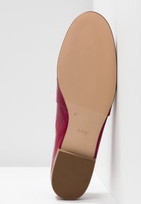Semelle d'une chaussure en cuir rouge avec une semelle extérieure texturée de couleur marron clair et un petit talon, reposant verticalement contre un mur blanc.
