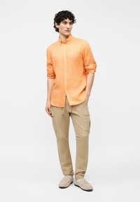 Άνδρας με σγουρά μαλλιά, φοράει πουκάμισο πορτοκαλί με κουμπιά, μανίκια ανασηκωμένα, μπεζ παντελόνι cargo και ανοιχτόχρωμα μοκασίνια, στέκεται σε λευκό φόντο.