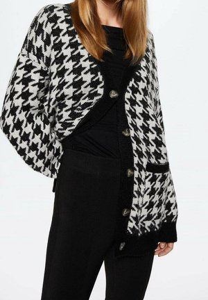 Personne portant un cardigan pied-de-poule noir et blanc avec bordure et boutons noirs, assorti à un haut et un pantalon noirs, haut du corps visible.