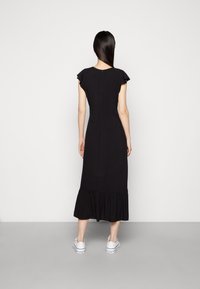 Nainen seisoo selkä kameralle päin, päällään musta midi-mekko, jossa on hulmuavat hihat, ja valkoiset tennarit, yksinkertaista taustaa vasten.