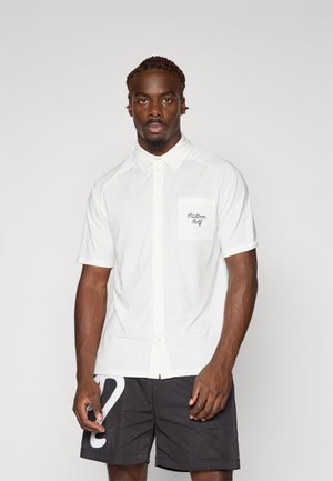 Hvit kortermet polo t-skjorte med teksturert finish, med en lomme som har "Malbon Golf" brodert med mørk tråd. Kombinert med svarte shorts.