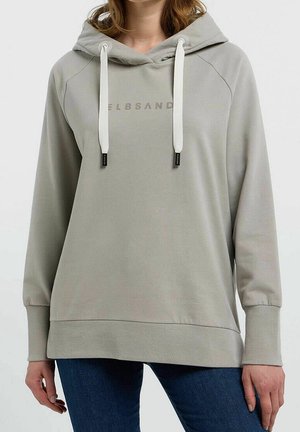 Mujer con sudadera con capucha gris claro con cordones blancos y texto tenue en el frente, combinada con vaqueros azul oscuro.