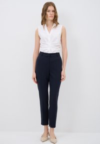 Blusa bianca senza maniche con scollatura a V e chiusura a bottoni, abbinata a pantaloni tailor navy a vita alta con vestibilità slim e spacco laterale.
