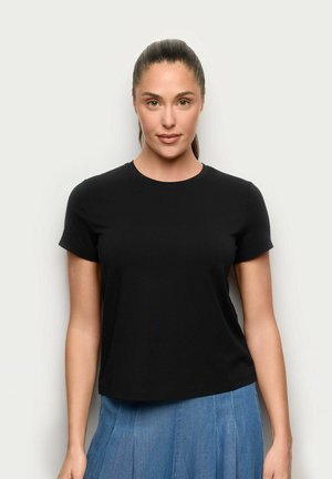 Kobieta z ciemnymi włosami związanymi do tyłu, ubrana w czarną koszulkę z krótkim rękawem i niebieską spódnicę, stojąca na tle gładkiego jasnego tła.