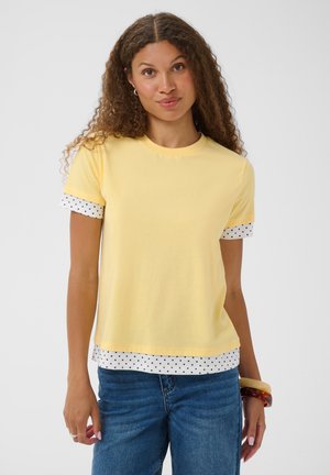 Femme aux cheveux bouclés portant un haut jaune à manches courtes superposé sur une chemise blanche à pois et un jean bleu, debout devant un fond uni.