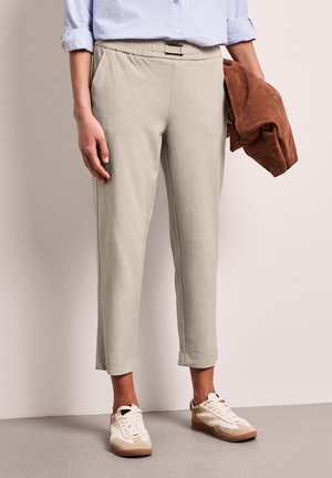 Persoon met beige cropped broek en witte sneakers, die een opgevouwen bruine jas vasthoudt, staand tegen een effen lichte muur.