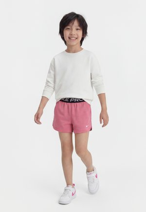 Smiling child walking, wearing white long-sleeve shirt, pink athletic shorts, and white sneakers with pink accents, against a plain white background.