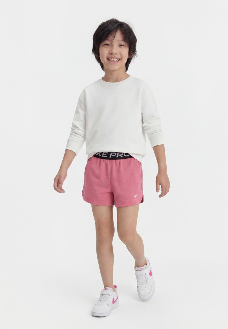 Χαμογελαστό παιδί που περπατά, φοράει λευκό μακρυμάνικο πουκάμισο, ροζ αθλητικά σορτς και λευκά παπούτσια με ροζ λεπτομέρειες, πάνω σε απλό λευκό φόντο.