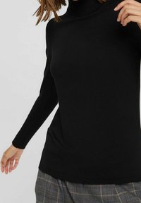 Zwarte coltrui met lange mouwen en een aansluitend ontwerp, gemaakt van zachte stof. Zichtbare geribbelde halslijn en zoom. Gecombineerd met geruite broek.
