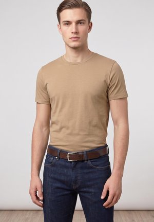 Young man wearing a beige short-sleeve t-shirt, dark blue jeans, and a brown leather belt standing against a plain gray background.