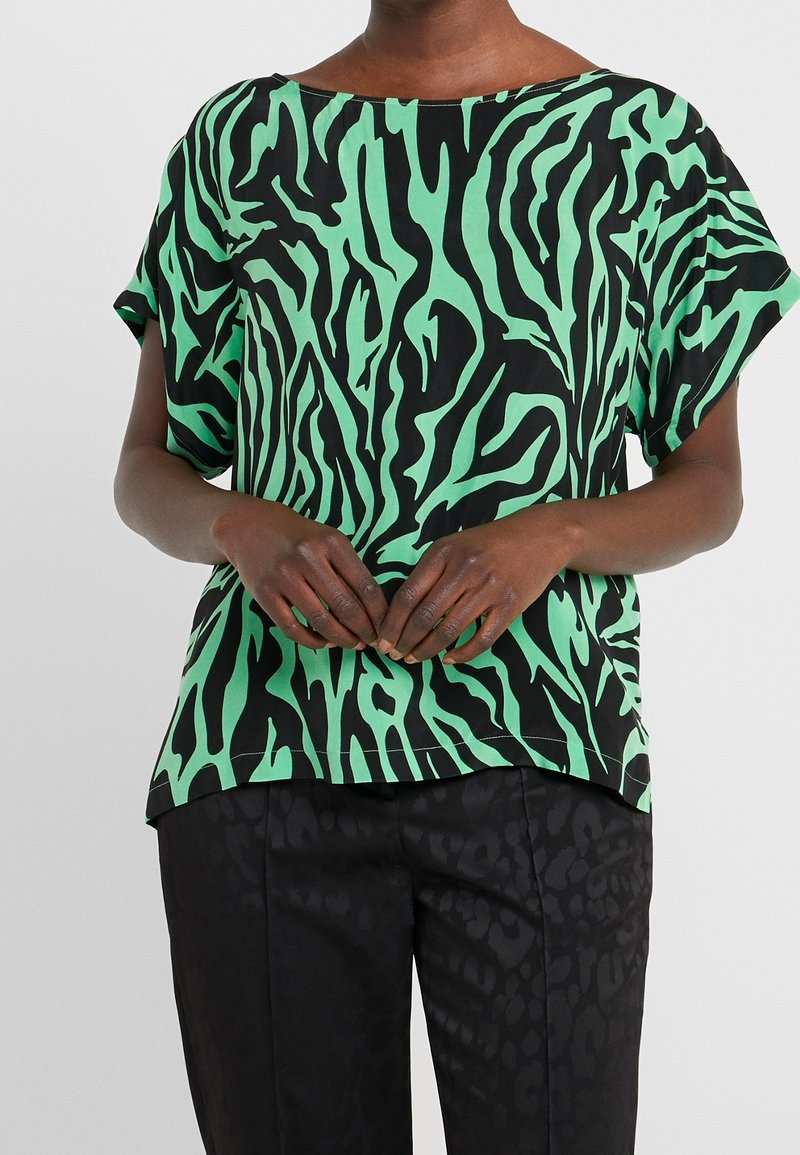 Svart topp med gröna zebramönster, korta ärmar och en bred halsringning. Tyget har en slät textur. Parat med svarta mönstrade byxor.