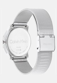 Sidabrinis nerūdijančio plieno laikrodis su tinkleliu, turintis apvalią dėžutę ir blizgiu dangteliu, su iškilniais ženklais ir vandeniui atsparus.