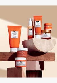 Sett med åtte oransje og hvite hudpleieprodukter arrangert på en lys bakgrunn, med varierende former og teksturer, hvor noen produkter støttes av treblokker.