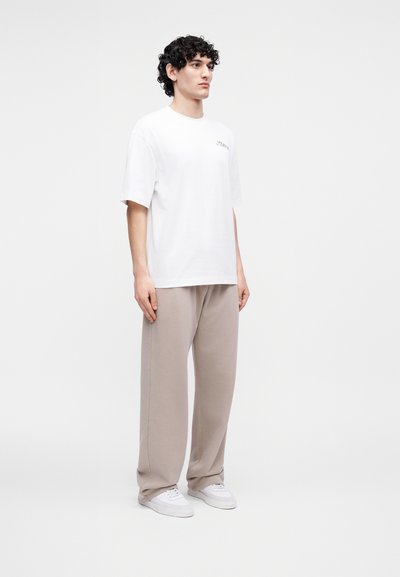 Νεαρός άνδρας με σγουρά μαλλιά, όρθιος, φορώντας λευκό φαρδύ μπλουζάκι, μπεζ άνετο παντελόνι και λευκά αθλητικά παπούτσια, μπροστά σε μονόχρωμο φόντο.