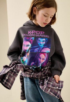 Dziecko noszące czarną bluzę z kapturem z trzema animowanymi postaciami i tekstem "K-POP DEMON HUNTERS,", trzymające fioletową koszulę w kratę zawiązaną wokół talii.