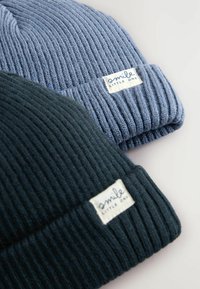 Deux bonnets en tricot côtelé, en bleu et en vert foncé. Chacun arbore une étiquette tissée avec "sourire petit" dans une couleur contrastante.