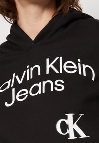 Sudadera negra de algodón, con un texto blanco en negrita "calvin Klein Jeans" y un logo blanco "ck" en el frente. Diseño clásico con capucha.