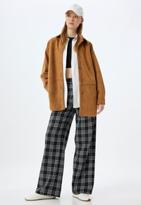 Veste surdimensionnée en suède marron, chemise blanche à boutons en dessous, pantalon large à carreaux noir et blanc, et baskets blanches.