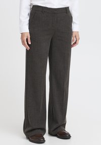Persoon in donkergrijze wijde broek, witte shirt en bruine leren schoenen, staande tegen een plain witte achtergrond.