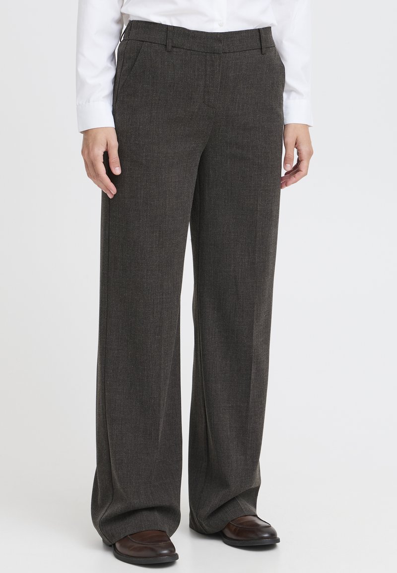 Persoon in donkergrijze wijde broek, witte shirt en bruine leren schoenen, staande tegen een plain witte achtergrond.