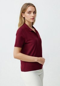 Burgundzka krótko rękawowa koszulka polo z prążkowanym dekoltem i teksturowanym materiałem. Model pozuje z boku, nosząc jasnobeżowe spodnie.