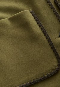 Gros plan sur un tissu en laine vert olive avec un détail en point de surjet marron foncé le long des bords d'une poche plaquée rectangulaire.