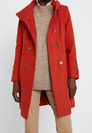 Kvinde iført en lys orange dobbeltradet frakke over en beige sweater og lysebrune bukser, stående mod en ensfarvet baggrund.