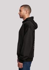 Black hooded sweatshirt with a relaxed fit, featuring a kangaroo pocket and ribbed cuffs. The back includes white printed text.