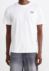 Man wearing a plain white short-sleeve t-shirt with a small sneaker patch on the left chest and faded gray jeans.