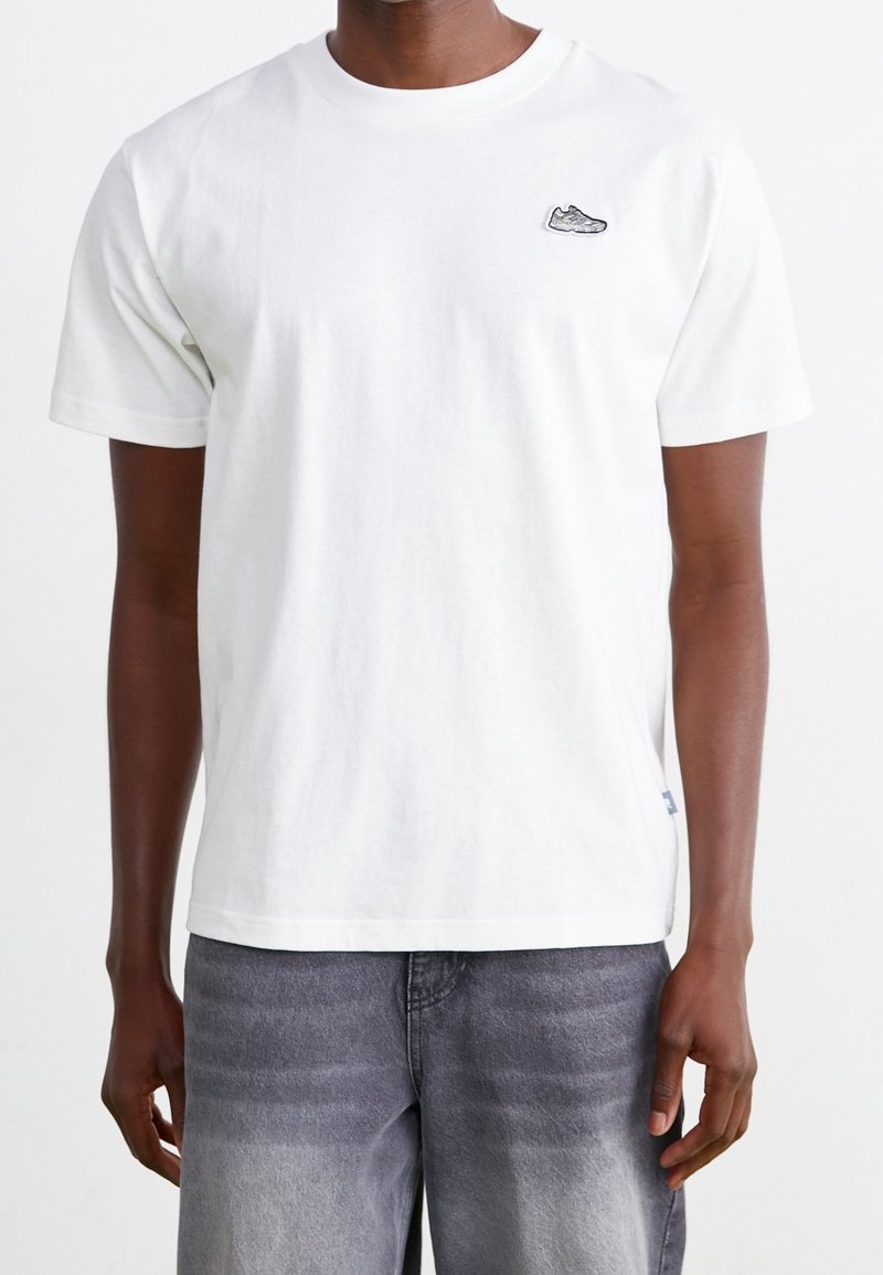 Man wearing a plain white short-sleeve t-shirt with a small sneaker patch on the left chest and faded gray jeans.