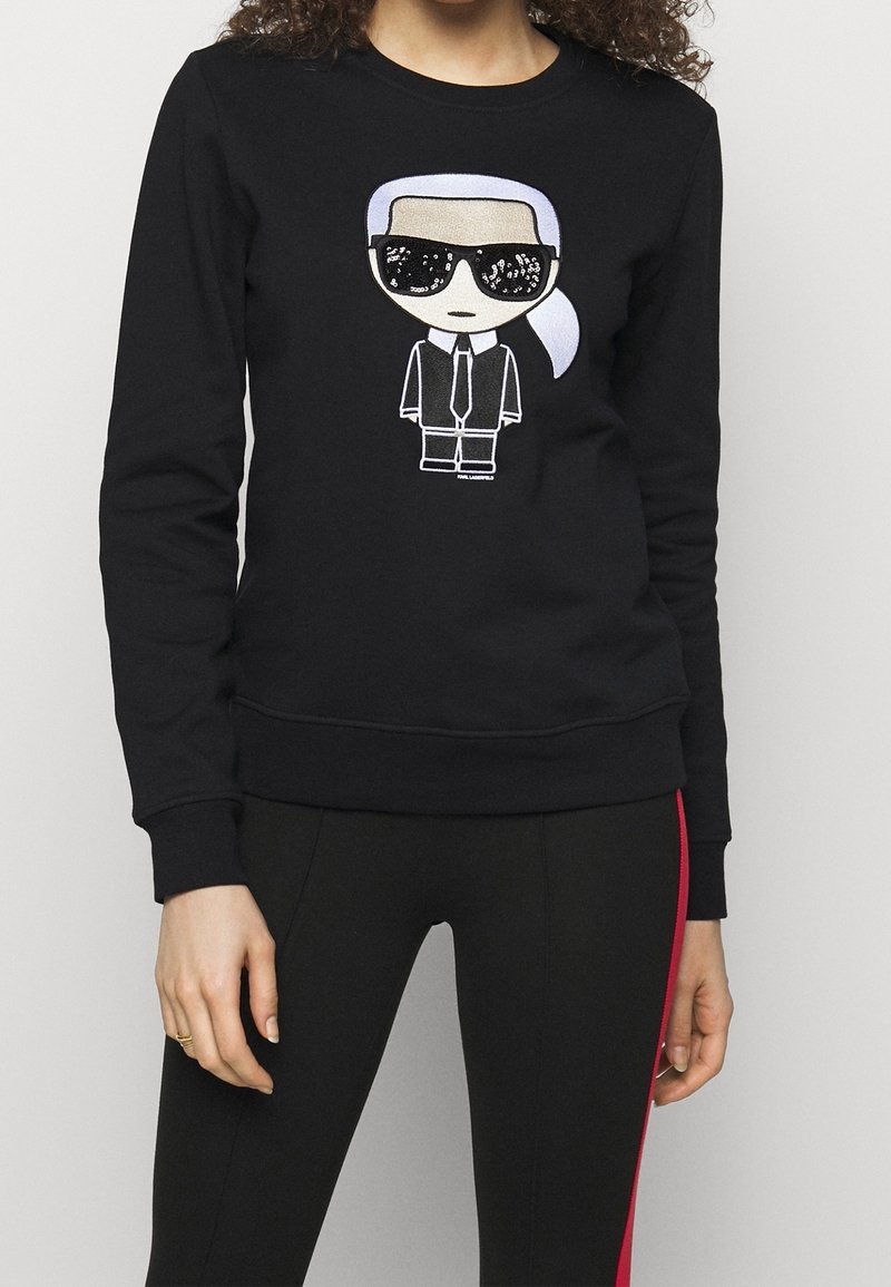 Personne portant un sweat-shirt noir avec une illustration de dessin animé représentant une silhouette aux lunettes de soleil, cheveux blancs attachés, costume noir et cravate sur le devant.