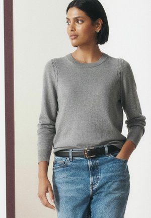 Kvinde med kort mørkt hår iført grå langærmet sweater og blå jeans med sort bælte, kigger til siden med den ene hånd i lommen.