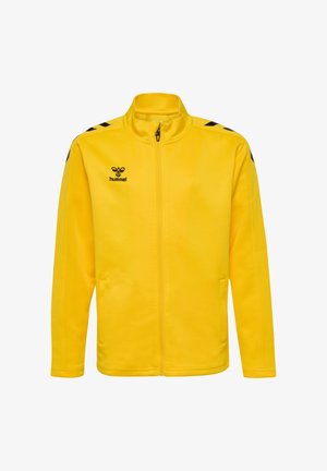 Smilende dreng iført en gul lynlåsjakke og sorte træningsbukser med sorte sneakers, stående mod en ensartet lys baggrund.