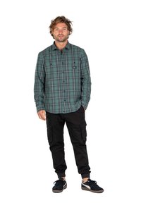 Camisa de franela a cuadros verdes y negros con cierre de botón, mangas largas, y pantalones cargo negros con bolsillos, combinados con zapatillas negras.