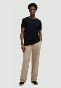 Schwarzes kurzärmliges T-Shirt mit dezentem Logo, kombiniert mit beigen weiten Hosen und schwarzen Schuhen, vor einem schlichten Hintergrund stehend.