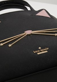 Black faux leather handbag with gold accents, featuring pink triangular ears, vertical gold whiskers, and a logo plate at the base.