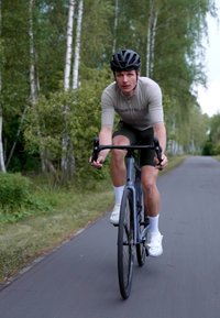 Un cycliste sur un vélo de route noir, portant un maillot gris et un short vert, des chaussettes blanches et des chaussures. Entouré d'arbres sur un chemin pavé.