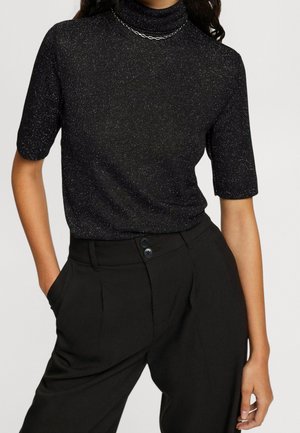 Persoon draagt een zwarte glinsterende korte mouwen coltrui, ingestopt in een zwarte broek met hoge taille, met handen in de zakken.