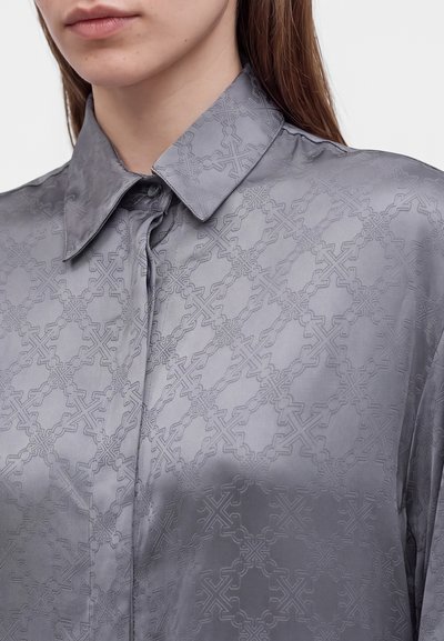 Gros plan d'une personne portant une chemise grise boutonnée avec un motif texturé géométrique subtil et un col pointu.