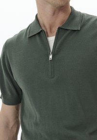 Polo de punto verde con mangas cortas, cuello con cremallera y tela texturizada. Presenta un tirador de cremallera plateado y un diseño ligero.